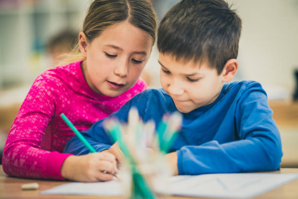 Girl holding her classmate's hand and guiding it to write in his notebook in classroom.