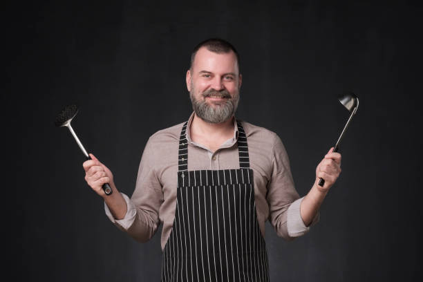 istock Mature european chef with beard holding a ladle standing on black studio wall. 1147519972