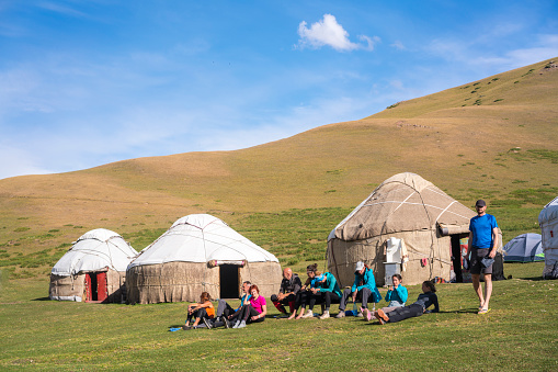 Tourists at camp resting on grassland, after day long horseback riding on the way to Song Kol Lake in Kyrgyzstan. Some are sitting, laying or standing while in background are yurts from local shepherds as camp facilities and tourists tent. It is moment of three days horseback riding, trekking over mountains from Kochkor to The Lake Song-Kul.