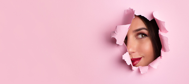 Beauty salon advertising banner with copy space. Beautiful girl looks through hole in pink paper background. Make up artist, fashion, beauty concept. Cosmetics sale