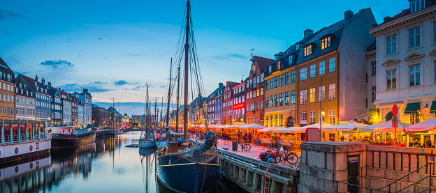 The iconic colourful restaurants and busy al fresco cafe bars of Nyhavn warmly illuminated beside the sailing ships moored beneath sunset skies in the heart of Copenhagen, Denmark’s vibrant capital city.