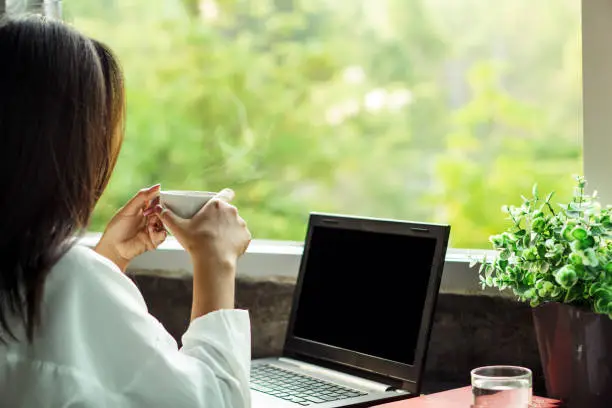 Photo of Asian woman drinking coffee at home office enjoying the view of nature from window