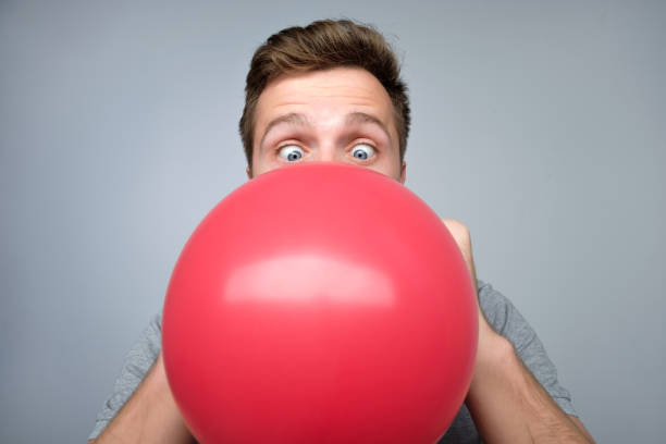 Young european man blowing up a red balloon Young european man blowing up a red balloon preparing for party on gray background inflating photos stock pictures, royalty-free photos & images