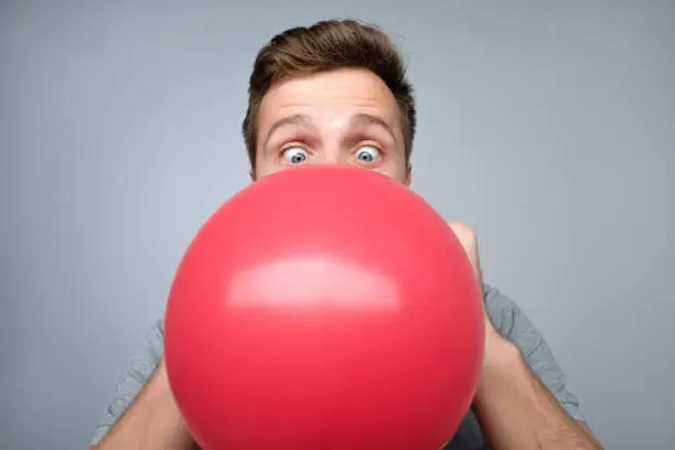 Young european man blowing up a red balloon preparing for party on gray background