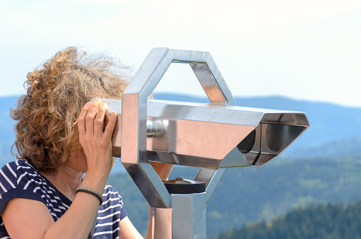 Woman tourist looking through a binocular telescope from a vantage point in mountainous terrain