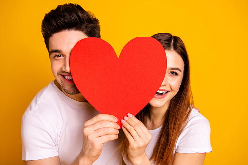 Close up photo beautiful she her he him his guy lady hiding facial expression laugh laughter hold hands arms heart shape paper postcard in love wear casual white t-shirts isolated yellow background