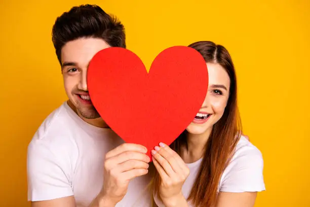 Photo of Close up photo beautiful she her he him his guy lady hiding facial expression laugh laughter hold hands arms heart shape paper postcard in love wear casual white t-shirts isolated yellow background