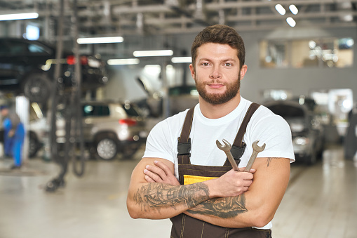 Handsome, muscular man wearing in white t shirt, coveralls, working as mechanic in autoservice. Brutal, tattoed repairman holding wrenches, posing, looking at camera.