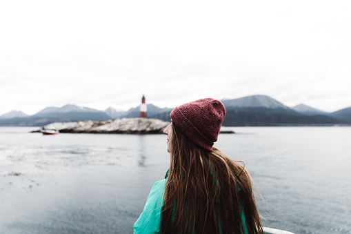 Woman in hat and green jacket sailing to the southernmost lighthouse in the world, looking at mountains and Pacific Ocean, Tierra Del Fuego region, Ushuaia, Argentina