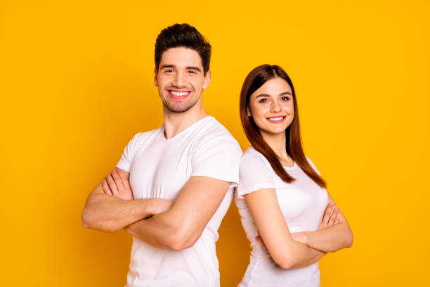 Close up photo beautiful she her he him his pair stand back side close teamwork self-confident professionals specialists work job reliable workers wear casual white t-shirts isolated yellow background