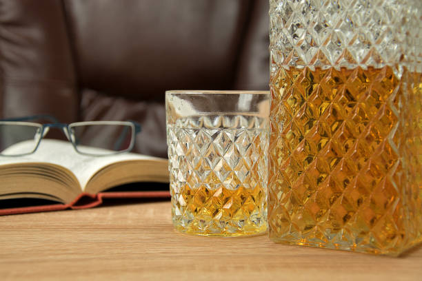 алкогольный напиток в кристаллическом декантере. в стакан налил виск�и на деревянный стол на заднем плане стул для сидения. открытая книга с - decanter crystal carafe glass стоковые фото и изображения