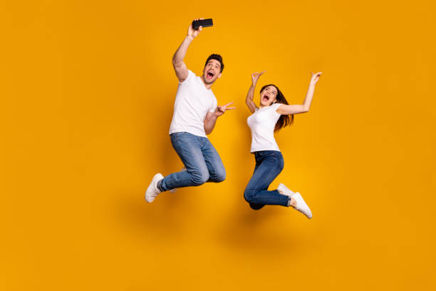 Full length side profile body size photo funky funny two people she her he him his guy lady jump high show v-sign make take selfies wear casual jeans denim white t-shirts isolated yellow background Full length side profile body size photo funky funny two people she her he him his guy lady jump high show v-sign make take selfies wear casual jeans denim white t-shirts isolated yellow background. man showing peace sign stock pictures, royalty-free photos & images