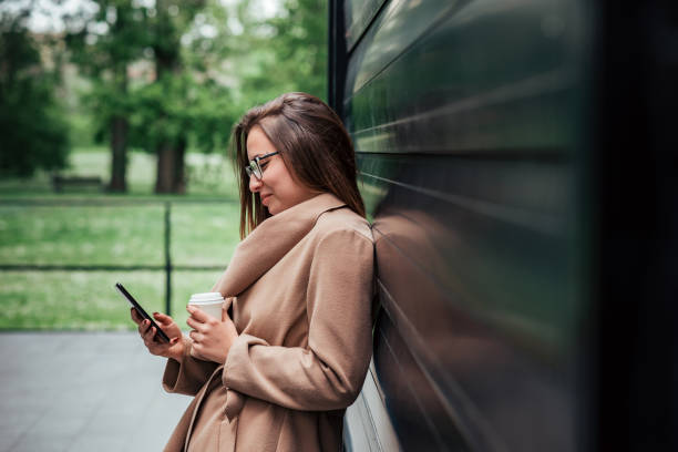 istock Beautiful smiling young woman with takeaway coffee using smart phone while leaning on a wall outdoors. 1147561246