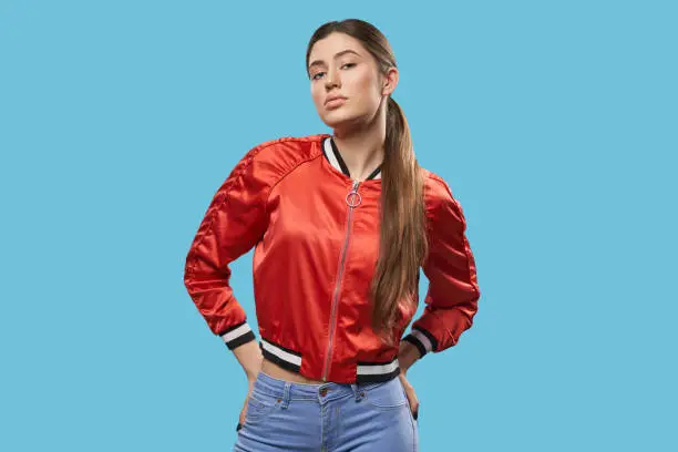 Portrait of young female model on blue background. Pretty girl with beautiful make up, long hair, ponytail, wearing in bright red bomber posing and looking at camera. Youth style.