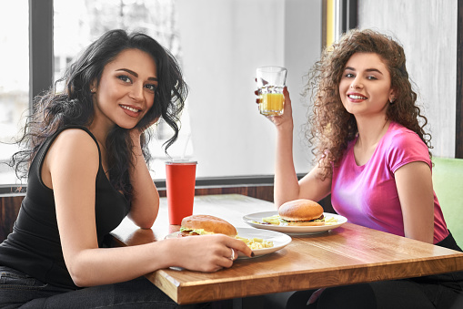 Two beautiful girls in t shirts sitting in cafe near window, posing and eating fast food, hamburgers. Pretty young women with curly hair and charming smile looking at camera, holding glass of juice.