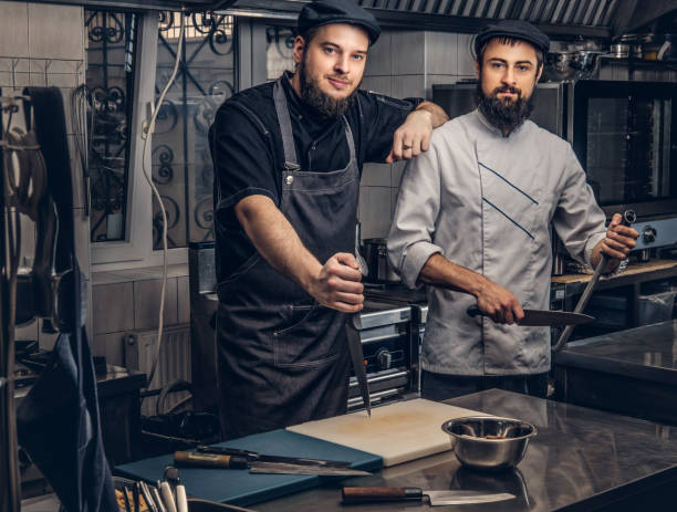 istock Two bearded cooks dressed in uniforms and hats posing in kitchen, looking at a camera. 1147563431