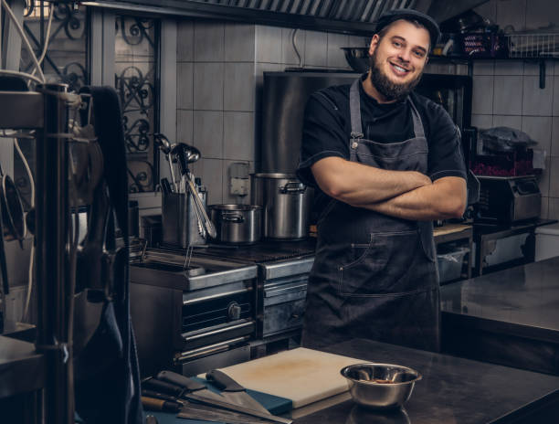 istock Smiling bearded cook in black uniform standing with crossed arms in kitchen. 1147564278