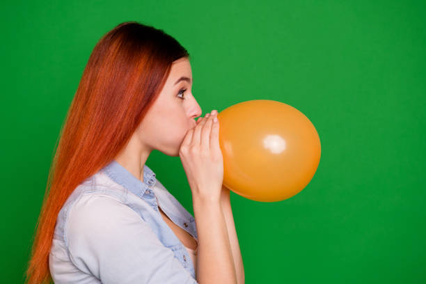 close up side profile photo beautiful amazing she her lady hold breath in big yellow air balloon arm hand weekend party holiday wear casual jeans denim shirt isolated green bright background - inflating balloon blowing air imagens e fotografias de stock