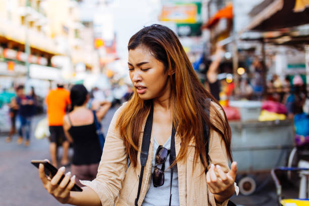 Traveling woman with broken phone on street Young Asian woman unhappy with badly functioning smartphone while traveling in city of Bangkok, Thailand poor signal stock pictures, royalty-free photos & images