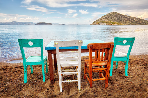Colorful retro wooden chairs and a table lined up on the beach at the seaside in Gumusluk, Bodrum, Turkey.