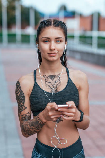 schöne frau lächelnd, fröhliches mädchen im sommer in der stadt, gebräunte hauttattoo. sportbekleidung top. telefon-ohrhörer. konzept der stärke, weiblichkeit, mut, zuversicht, gedanken der inspiration, motivation. - headphones music mobile phone tattoo stock-fotos und bilder