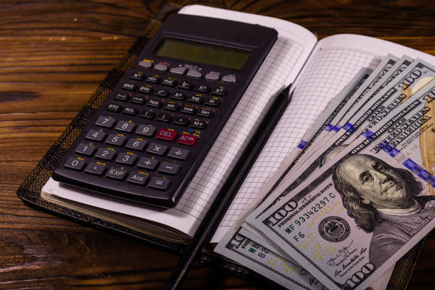 istock Opened notepad, one hundred dollar banknotes, scientific calculator and pencil on wooden table 1147653219