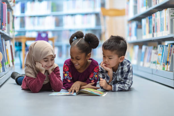 Children Reading Three (3) kids who are friends are laying on the floor in between bookshelves in the library. They are reading a book together. They are all smiling and enjoying their afternoon. modest clothing stock pictures, royalty-free photos & images