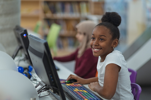 A young girl of African descent smiles as she sits at a computer in the library. She is smiling off into the distance while others focus on their own screens.