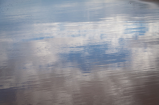 Blue sky and clouds reflected off wet sand on a beach. Water surface with sky reflection summer texture, background