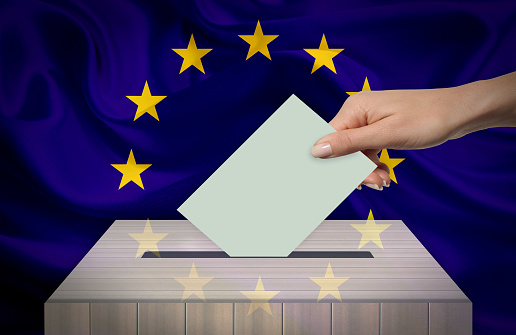 Hand with voting ballot and box in front of the EUROPEAN UNION flag.