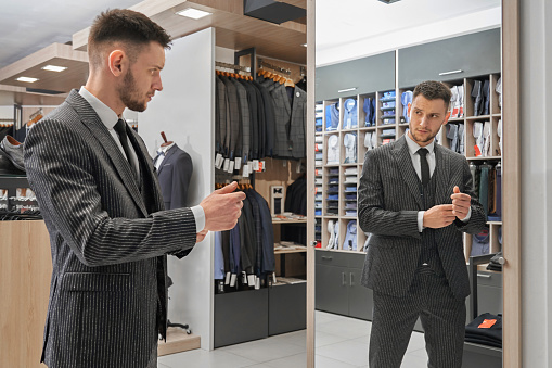 Handsome man in elegant striped suit standing, looking at mirror in boutique and choosing clothing. Young client in fashionable jacket, white shirt and black necktie.