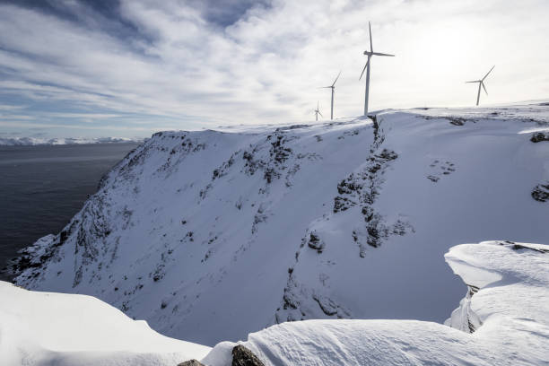 Aerial view of a wind farm in a snow-covered landscape at sunset. Wind turbines stand tall against the warm glow of the setting sun, creating a contrast between renewable energy and the winter scenery.