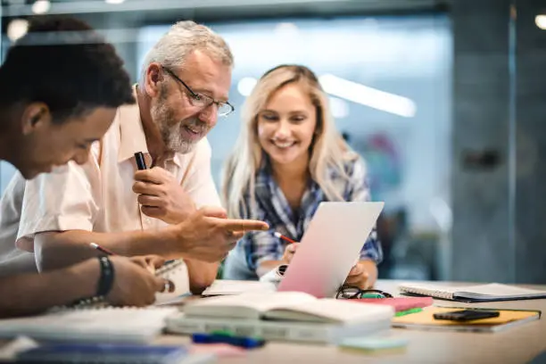 Photo of Happy mature professor and his students using laptop at campus.