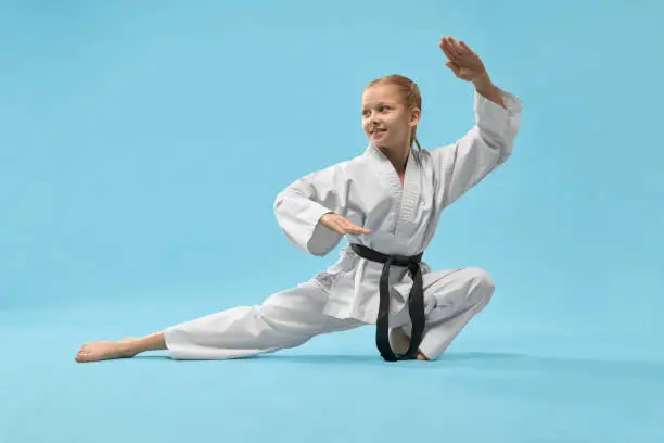 Cheerful little sports woman sitting on twine and practising karate on blue isolated background. Active girl wearing white kimono and black belt doing sport in studio. Concept of karate and jujitsu.