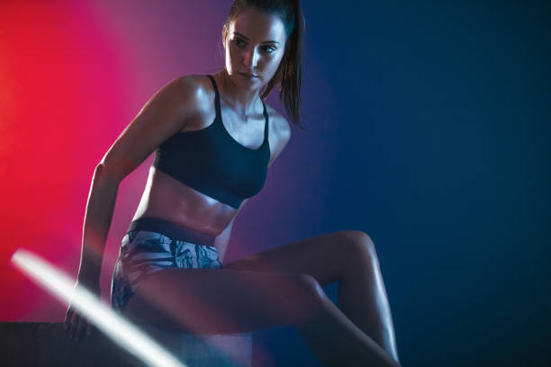 She is getting ready for exercise Female athlete sitting on a box over colored background with gel effect. Sportswoman getting ready for exercising. crossfit girl stock pictures, royalty-free photos & images