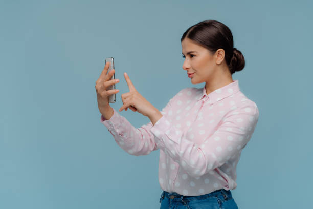 o tiro lateralmente da senhora triguenha tem o cabelo penteado, prende o telefone de pilha moderno, faz o retrato do selfie, desgasta a camisa e as calças de brim do ponto polka, isoladas sobre o fundo azul. mulher de negócios que fotografa ela mesma - coque - fotografias e filmes do acervo