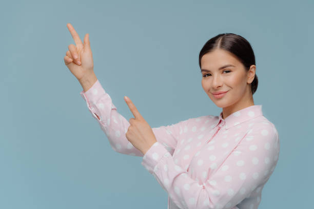 istock Smiling Caucasian female points straight to top, demonstrates something at upper left corner, has pleased facial expression, wears polka dot shirt, isolated over blue background. Lady points up 1147782793