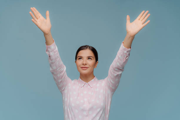 Cheerful lovely brunette female tries to catch something falling from sky, stretches hands focused upwards, shows palms, dressed in polka dot shirt, poses over blue background. Come to me closer!
