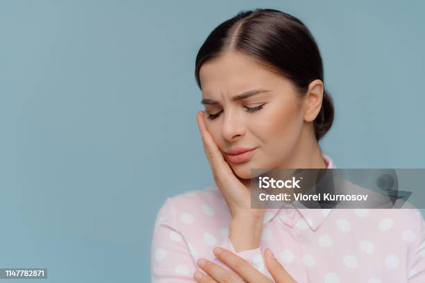 Brunette Woman Suffers From Terrible Strong Teeth Pain Touches Cheek With Hand Needs To Dentist Closes Eyes From Pain Dressed In Polka Dot Shirt Isolated On Blue Studio Wall Dental Care Concept Stock Photo - Download Image Now