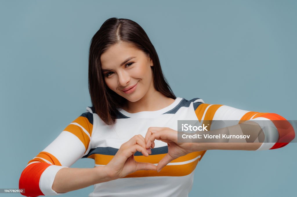 Nice looking brunette woman shows heart gesture, wears casual clothes, expresses love and sympathy, models over blue background. Romantic female demonstrates valentine with hands, stands indoor Adult Stock Photo