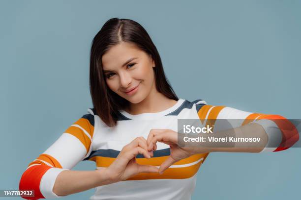 Nice Looking Brunette Woman Shows Heart Gesture Wears Casual Clothes Expresses Love And Sympathy Models Over Blue Background Romantic Female Demonstrates Valentine With Hands Stands Indoor Stock Photo - Download Image Now