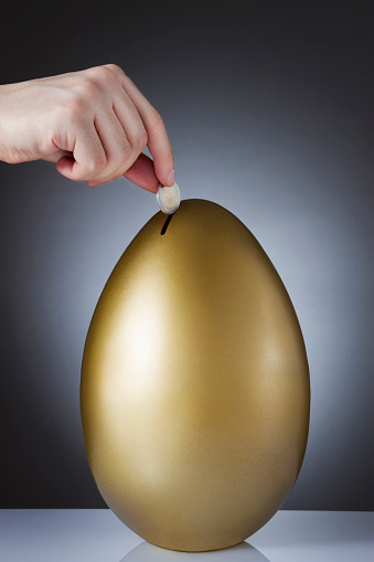 Saving concept with human hand gold colored giant egg coin bank on dark gray background with copy space