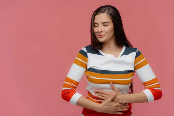 Satisfied woman keeps hands on belly, feels replete after delicious supper, has full stomach, long hair, appealing appearance, wears jumper, models on pink wall. Lady finds out about pregnancy