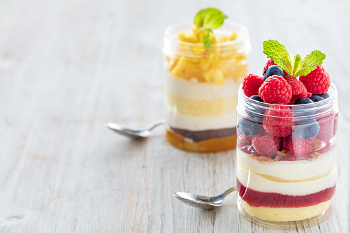 Raspberry, blueberries  and mango dessert, cheesecake, trifle, mouse in a glass jar with small spoons  on a light wooden background