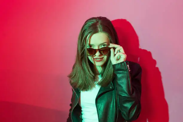 Photo of Fashioned girl in leather jacket in dark glasses in pink and blue  neon lights background.