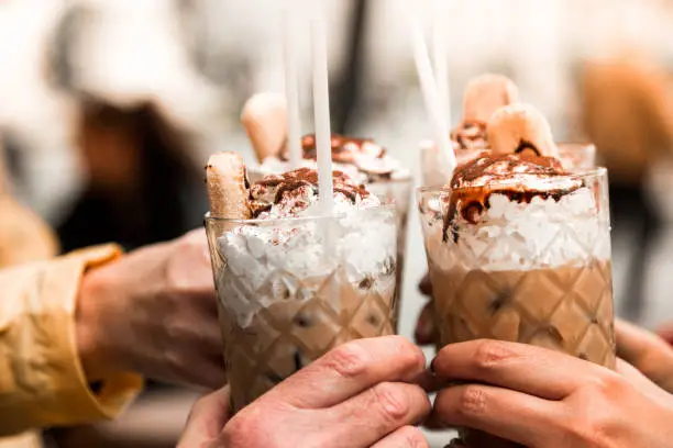 Close up color image depicting people celebrating with glasses of iced coffee at an outdoors cafe in summer. The people are celebrating with a toast and are touching their glasses together in a celebration gesture. The people are unrecognisable and we can only see their hands. The background of a city centre is totally defocused, leaving room for copy space.