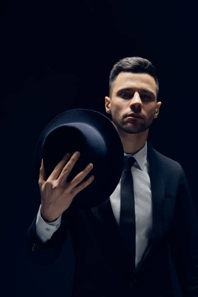 istock Young handsome man in black suit wearing his hat on dark background 1147802315