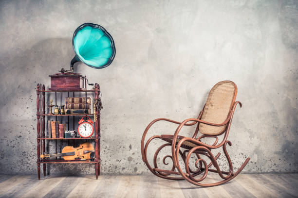 istock Vintage antique gramophone phonograph with aquamarine horn, books, clock, camera, binoculars, fiddle, keys on shelf, aged rocking chair front concrete wall background. Retro old style filtered photo 1147846516