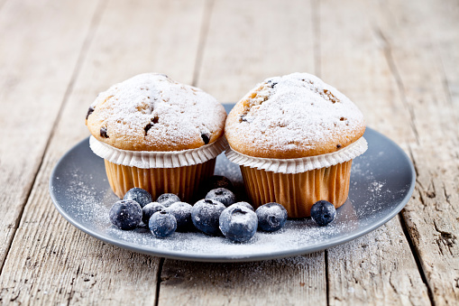 Homemade fresh muffins with sugar powder and blueberries on ceramic plate on rustic wooden table background.