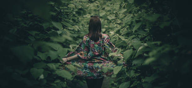 Woman in a gown with flowers posing in a lush green jungle like forest or bush. Leaves surrounding a cute redhead girl exposing her colorful dress. Mystical photo of a girl in a leafy forest.